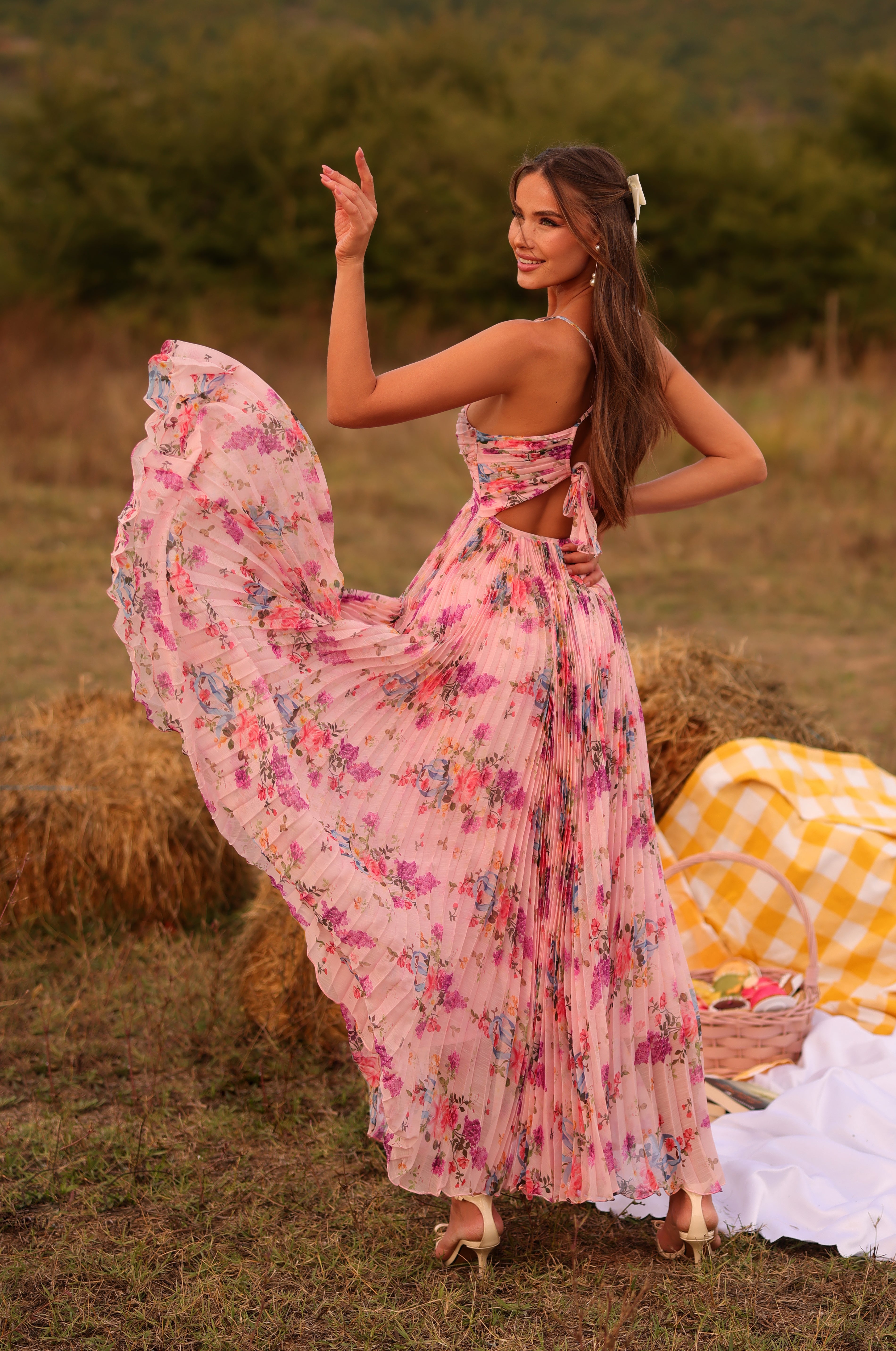 A woman in the Pleated Blush Pink Floral Maxi Dress: Florinne poses outdoors, lifting her skirt near hay bales and a yellow-checkered picnic blanket.